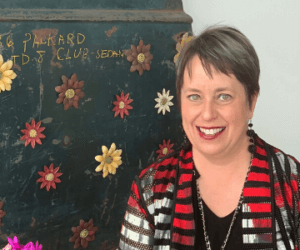 A woman with short brown hair smiles at the camera. She's wearing a black top and a red-and-black patterned jacket. Behind her is a dark chalkboard-style wall painted with colorful daisies and faded lettering.
