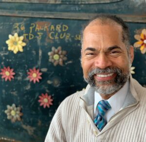 Man smiling at the camera, wearing a cream ribbed quarter-zip sweater over a white shirt and blue striped tie, seated in front of a weathered dark green vintage truck door decorated with painted flowers and the hand-lettered words '36 Packard.'