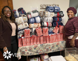 2 POWER volunteers pose in front of a large pile of holiday gifts and food they are preparing to distribute.