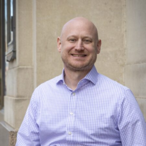 Smiling bald man with a short reddish beard, wearing a lavender gingham button-down shirt, standing outdoors against a pale stone building.
